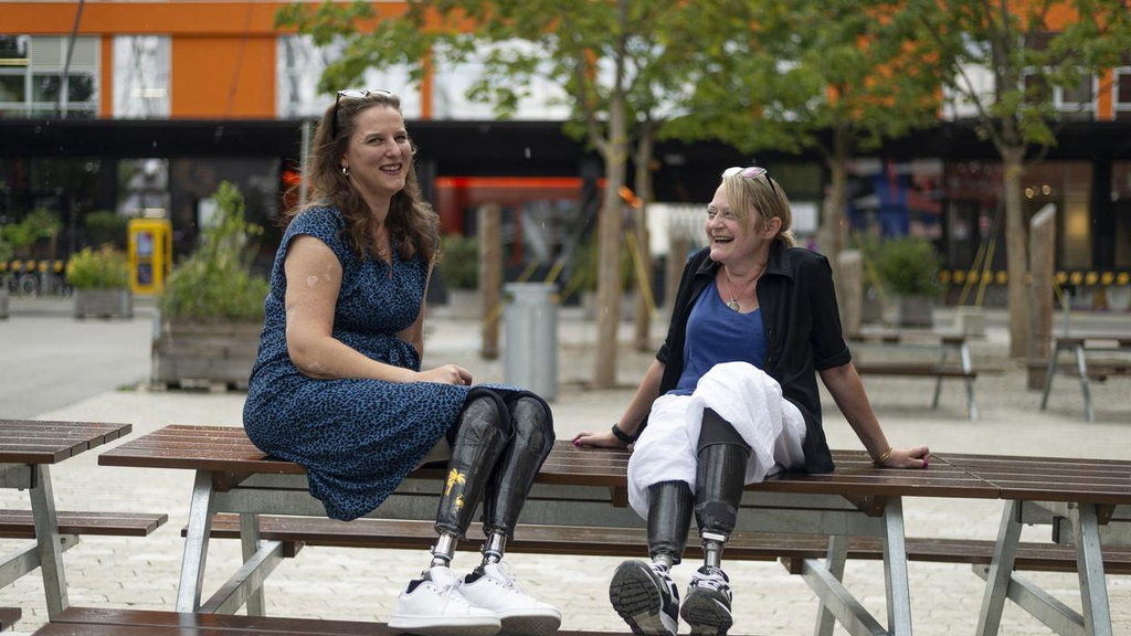 Zwei Frauen sitzen gemeinsam auf dem Tisch einer Picknickbank auf einem großen Platz. Beide Frauen haben zwei Unterschenkelprothesen.