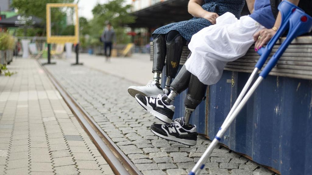 Am rechten Bildrand sitzen zwei Frauen, deren Unterschenkelprothesen über den Rand herabbaumeln. Im Vordergrund stehen zwei blaue Krücken.