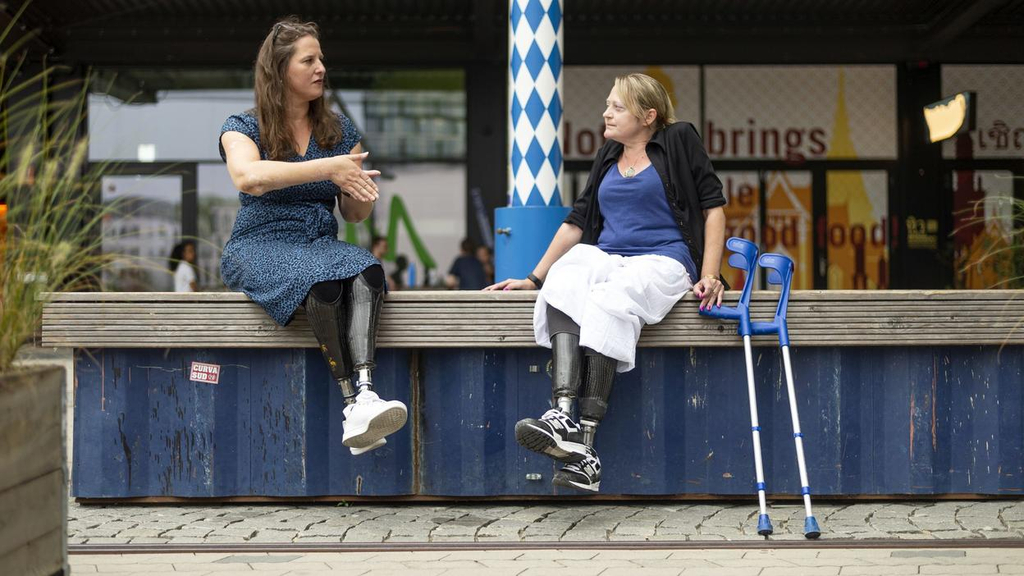 Auf einem Podest sitzen zwei Frauen mit Unterschenkelprothesen und unterhalten sich. Eine der beiden Frauen scheint gerade etwas zu erklären.