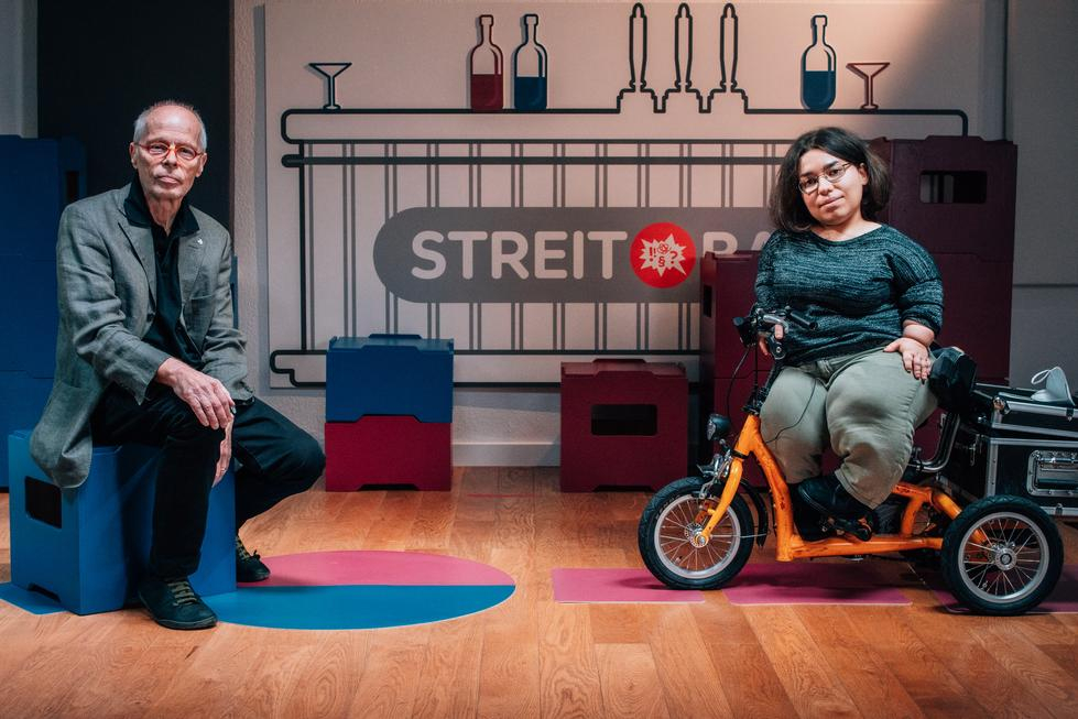 Ein Mann in höherem Erwachsenenalter und eine junge, kleinwüchsige Frau sitzen nebeneinander in einem Studio