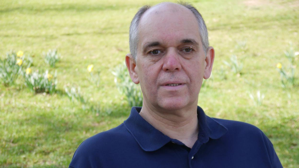 Ein Mann im blauen Polohemd steht vor einer grünen Wiese und schaut in die Kamera.