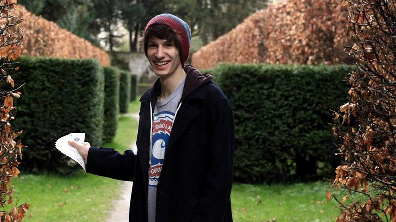 Jugendlicher steht mit Mantel und Mütze vor Hecken