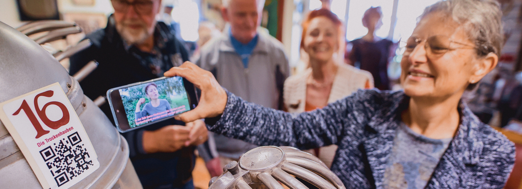 Eine Gruppe Besucher*innen steht an einem alten Frisierstuhl. Über ein Mobiltelefon schauen sie sich ein Gebärdensprachvideo zum Exponat an.