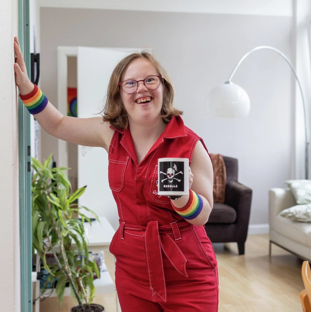 Natalie Dedreux steht in einem roten Jumpsuite in einem Wohnzimmer und lacht in die Kamera. Sie hält sich mit der rechten Hand an der Wand fest.