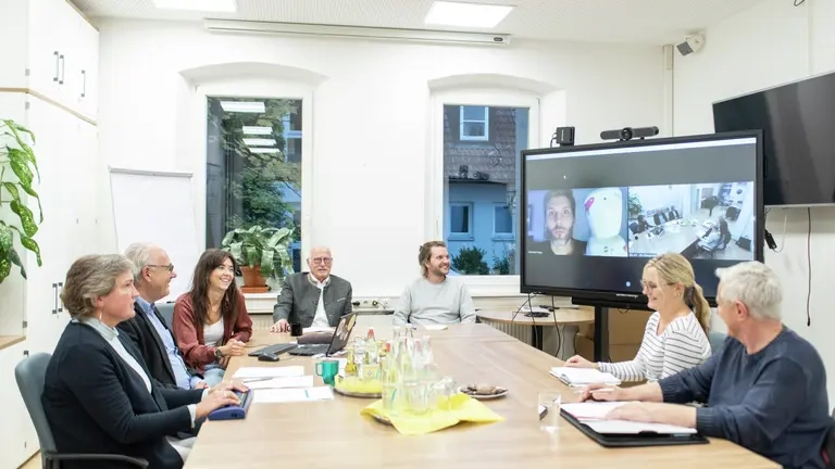 Eine Gruppe von Menschen sitzen in einer Konferenz, einige nehmen per Videokonferenz teil