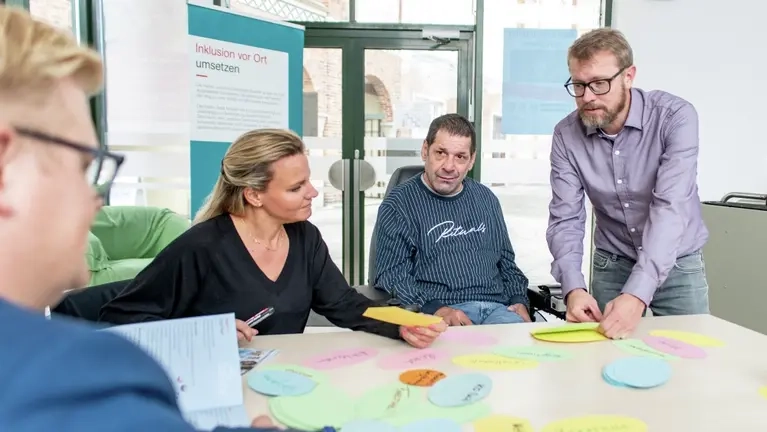 Menschen mit und ohne Behinderung sitzen an einem Tisch und schreiben sich Stichwörter auf ovale, bunte Kärtchen auf