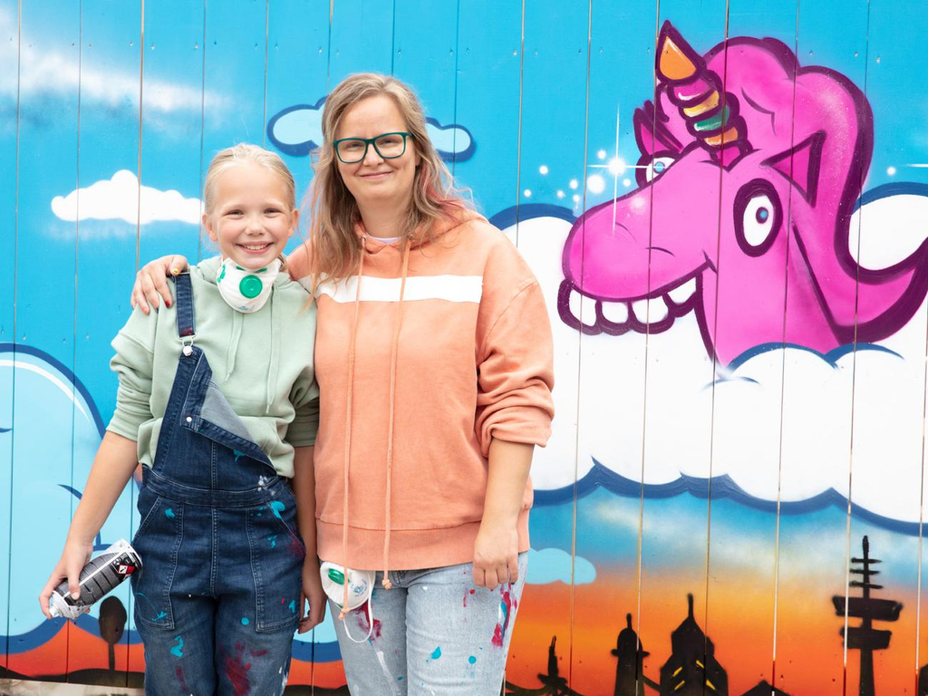Ein Mädchen und eine Frau stehen Arm in Arm nebeneinander. Sie tragen farbbespritzte Kleidung. Sie stehen vor einer Wand mit einem Einhorn-Graffiti.
