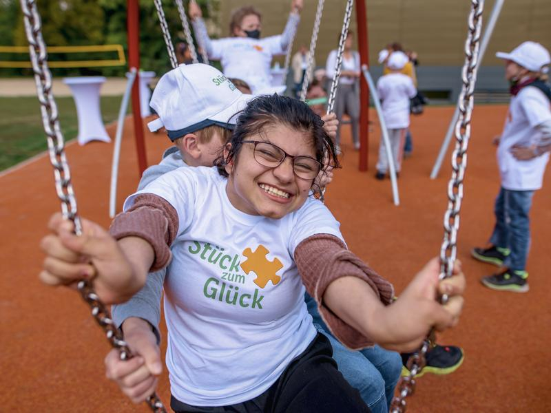 Ein Mädchen sitzt lachend auf einer Schaukel. Sie trägt ein T-Shirt mit dem Aufdruck Stück zum Glück.