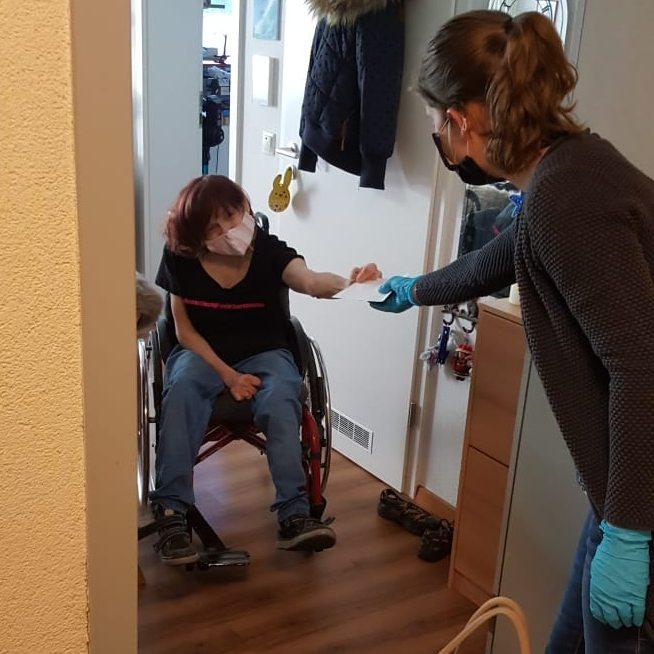 Eine Frau im Rollstuhl an ihrer Wohnungstür, die Einkäufe von einer anderen Frau annimmt. Beide Frauen tragen einen Mundschutz.