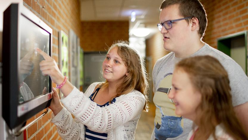 Zwei Mädchen und ein Junge stehen in einem Schulflur und schauen auf einen Monitor. Ein Mädchen zeigt auf den Monitor.