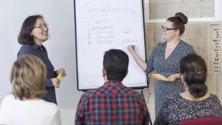Zwei Frauen zeigen auf Stichworte auf einem Flippchart, mehrere Menschen schauen zu