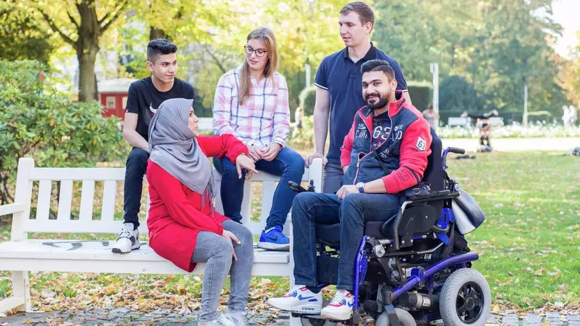 Eine Gruppe von Menschen sitzen im Park, eine Person sitzt im Rollstuhl, eine trägt ein Kopftuch.