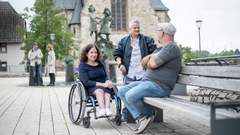 Drei Personen, eine sitzt im Rollstuhl, reden miteinander