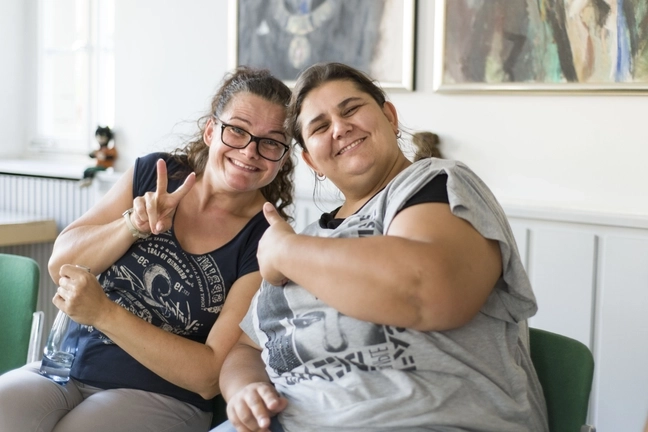 Zwei Frauen lachen in die Kamera, eine zeigt "Daumen hoch", die andere macht ein "Peace"-Zeichen.