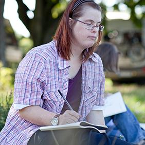 Junge Frau mit langen Haaren und Brille sitzt im Gras und schreibt etwas.