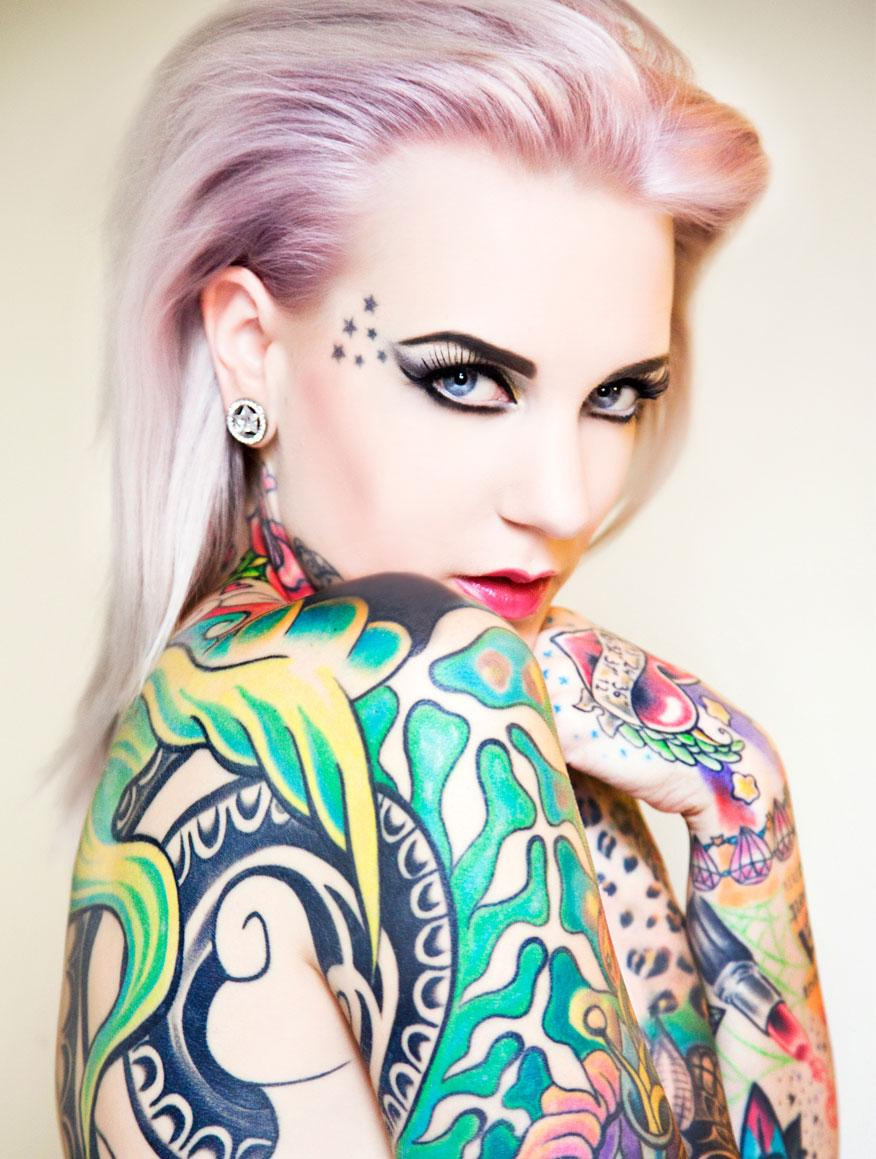 Foto einer im Arm – und Schulterbereich über und über bunt tätowierten, stark geschminkten jungen Frau.