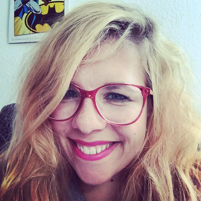 Eine junge Frau mit langen blonden Haaren und roter Brille lächelt in die Kamera