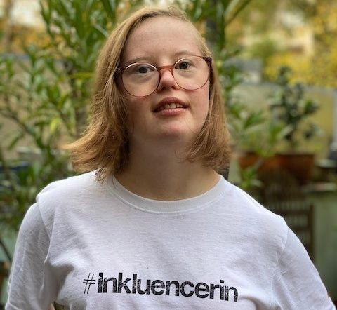 Porträt einer jungen Frau mit Down-Syndrom. Sie trägt ein T-Shirt mit der Aufschrift: #Inkluencerin