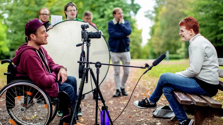 Ein junger Mann im Rollstuhl interviewt eine Frau in einem Park. Eine Kamera mit Stativ und Mikrofon sind auf die Frau gerichtet.