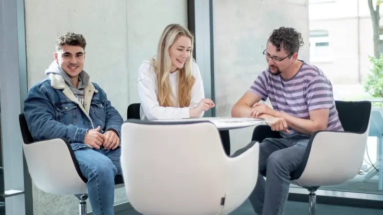 Drei junge Menschen sitzen an einem Tisch und schauen sich ein Dokument an.