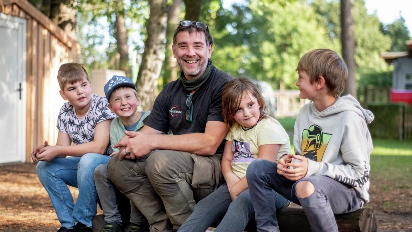 Vier Kinder und ein Erwachsener sitzen auf einem Bank aus einem Baumstamm in einem Park