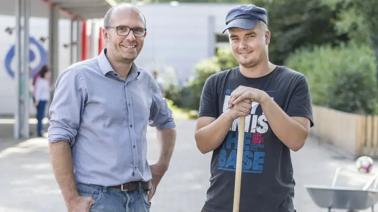 Zwei Männer: einer mit einer Schaufel mit Capi und T-shirt, der andere mit Hemd, beide schauen in die Kamera