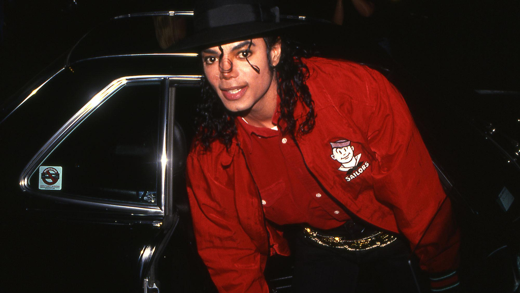 Michael Jackson mit Hut und roter Jacke vor einem schwarzen Auto