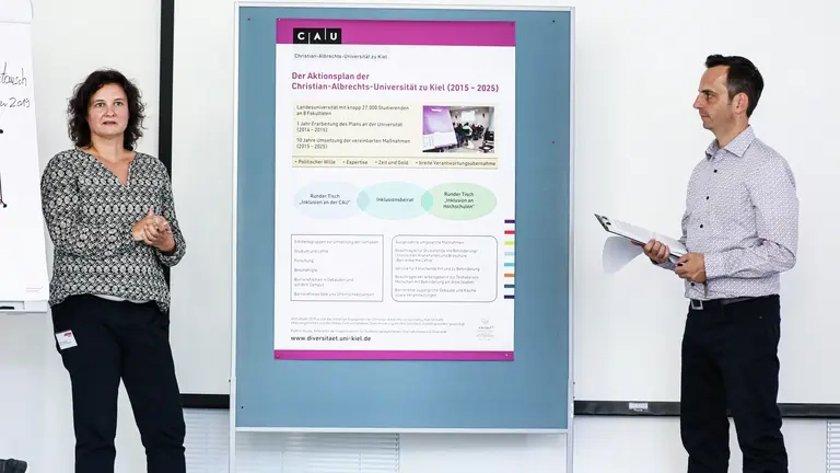 Eine Frau und ein Mann stehen an einer Pinnwand mit Grafik und erklären sie.