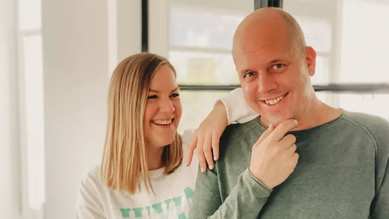 Ralph Ruthe und seine Frau Tina. Sie hat ihren Arm auf seine Schulter gelegt und lacht ihn an. Er schaut verschmitzt in die Kamera.