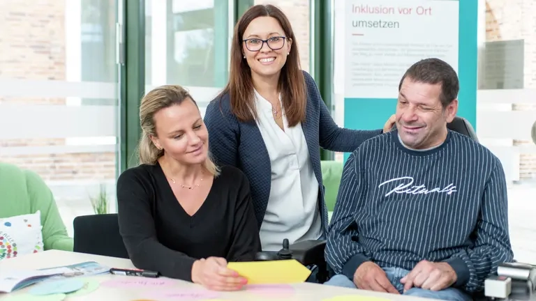 Eine Frau steht bei einem Mann im Rollstuhl und einer Frau, die sich eine Karteikarte ansehen.