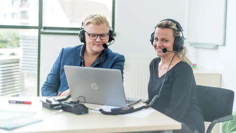 Ein Mann und eine Frau sitzen mit Head-Set vor eine Laptop.