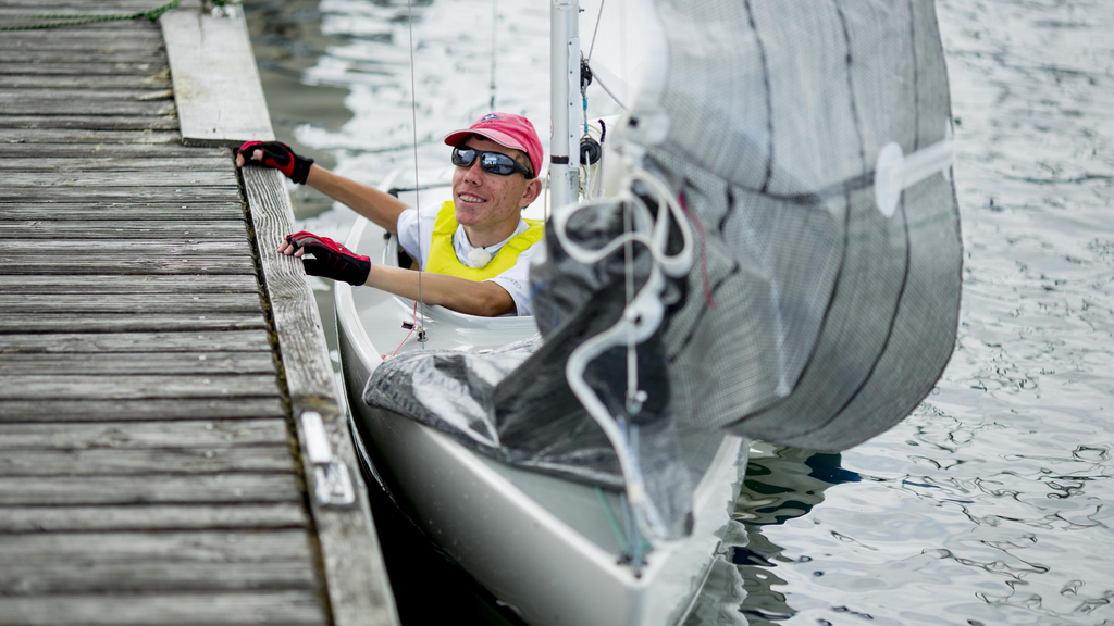 Ein junger Mann mit Behinderung sitz in einem Segelboot und hält sich am Bootssteg fest.