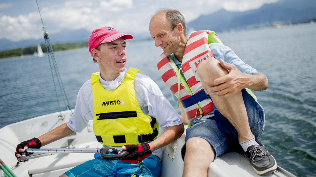 Ein junger Mann mit Behinderung und ein Mann sitzen nebeneinander auf einem Segelboot.