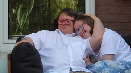 Ein junges Pärchen - beide mit Downsyndrom - sitzen auf dem Sofa. Er hat seinen Arm um sie gelegt und sie kuschelt sich an ihn an.