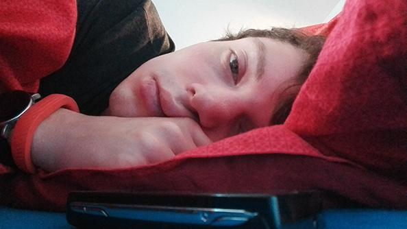 Junger Mann liegt mit müden geöffneten Augen im Bett, neben sich ein Handy.