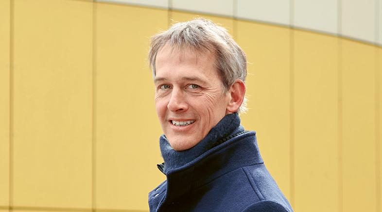 Ein Mann im dunkelblauen Mantel steht vor einem gelben Gebäude