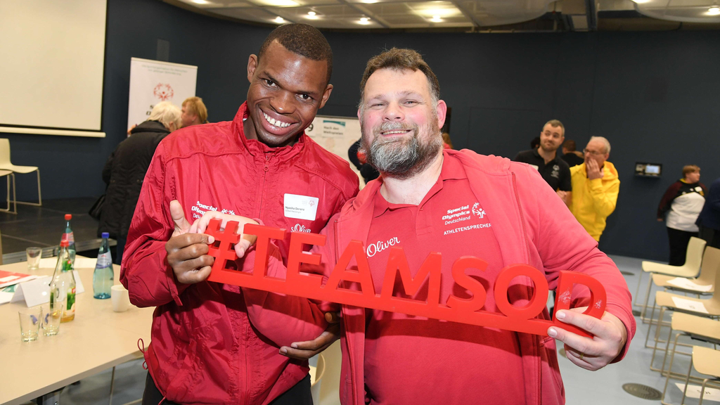 Ein junger schwarzer Mann und ein weißer Mann mittleren Alters stehen lächelnd nebeneinander und halten den Schriftzug #TEAMSOD fest