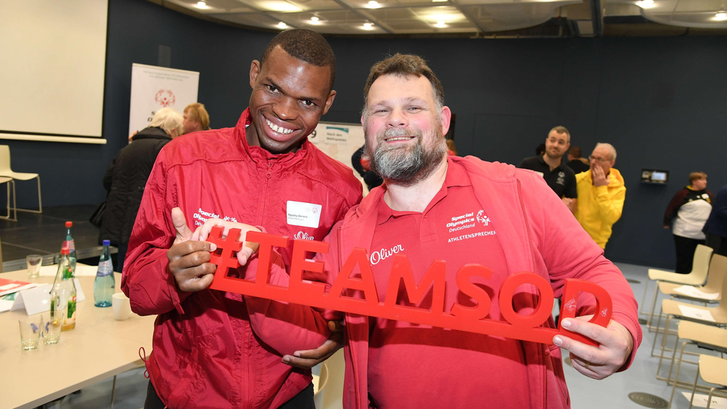 Ein junger schwarzer Mann und ein weißer Mann mittleren Alters stehen lächelnd nebeneinander und halten den Schriftzug #TEAMSOD fest