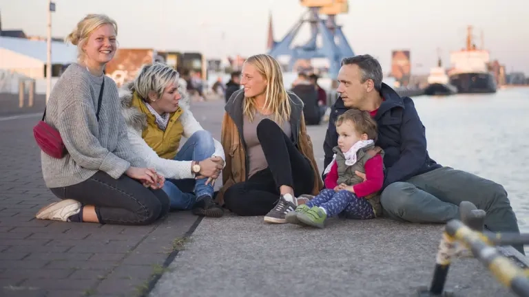 Eine Familie mit drei Kindern sitzt am Stadthafen, im Hintergrund sind Schiffe, Kräne und das Meer zu sehen