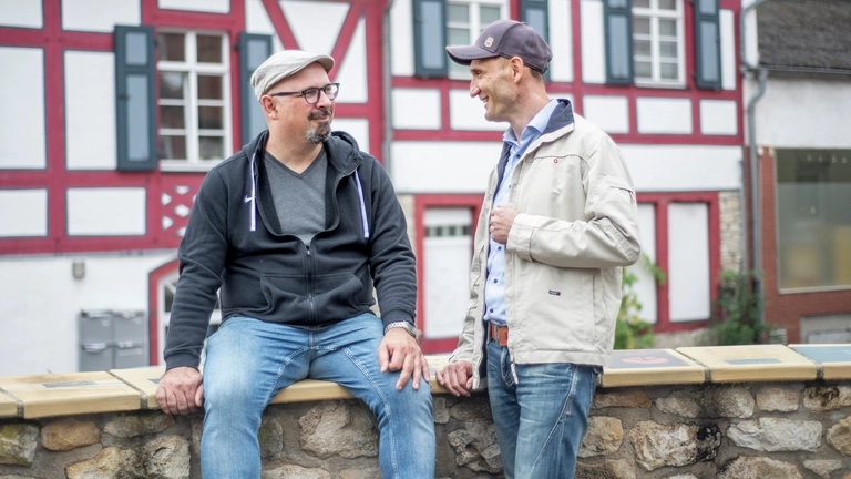 Zwei Männer unterhalten sich, einer sitzt auf einer kleinen Mauer, im Hintergrund sind Fachwerkhäuser.