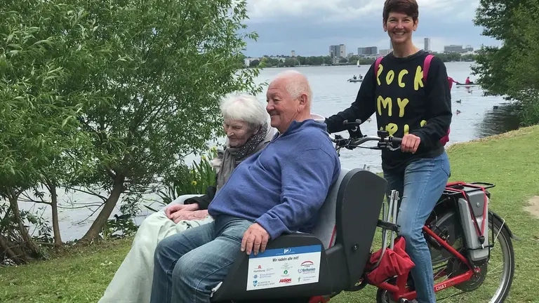 Eine lächelnde junge Frau fährt ein älteres Paar (Mann und Frau) am Ufer eines Gewässers in einer Rikscha spazieren.