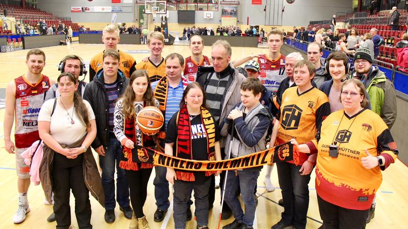 Gruppenbild: Basketballsfans, teils mit sichtbarer Sehbehinderung, Spieler  und weitere Personen in einer Halle