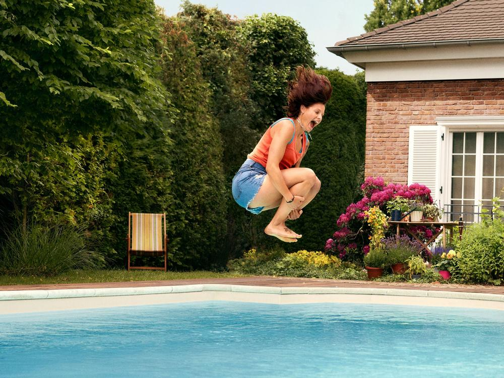 Eine junge Frau in Shorts und Top macht lachend eine Arschbombe in ihren Pool.