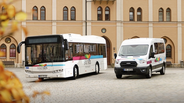 Ein großer Stadtbus und ein kleinerer Sprinter-Expressbus stehen auf einem Kirchevorplatz.