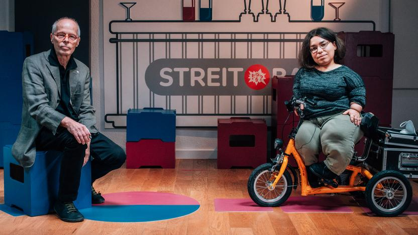 Ein Mann in höherem Erwachsenenalter und eine junge, kleinwüchsige Frau sitzen nebeneinander in einem Studio