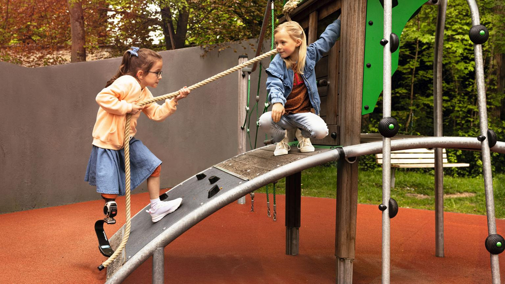 Zwei Mädchen spielen auf einem barrierefreien Spielturm. Ein Mädchen mit Beinprothese erklimmt den Turm gerade, indem sie sich an einem Seil über eine Rampe hochzieht.