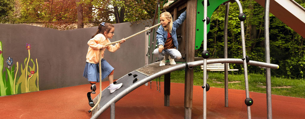 Zwei Mädchen spielen auf einem barrierefreien Spielturm. Ein Mädchen mit Beinprothese erklimmt den Turm gerade, indem sie sich an einem Seil über eine Rampe hochzieht.