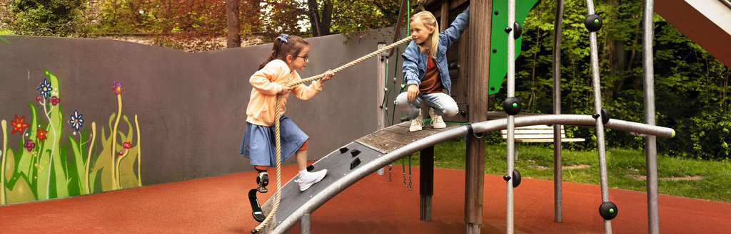 Zwei Mädchen spielen auf einem barrierefreien Spielturm. Ein Mädchen mit Beinprothese erklimmt den Turm gerade, indem sie sich an einem Seil über eine Rampe hochzieht.