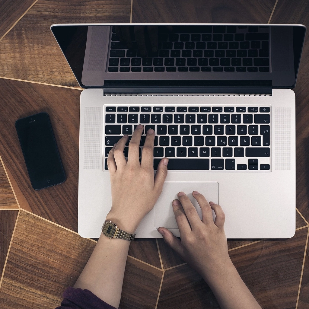 Eine Person tippt an einem Laptop, neben dem Laptop liegt ein Smartphone.