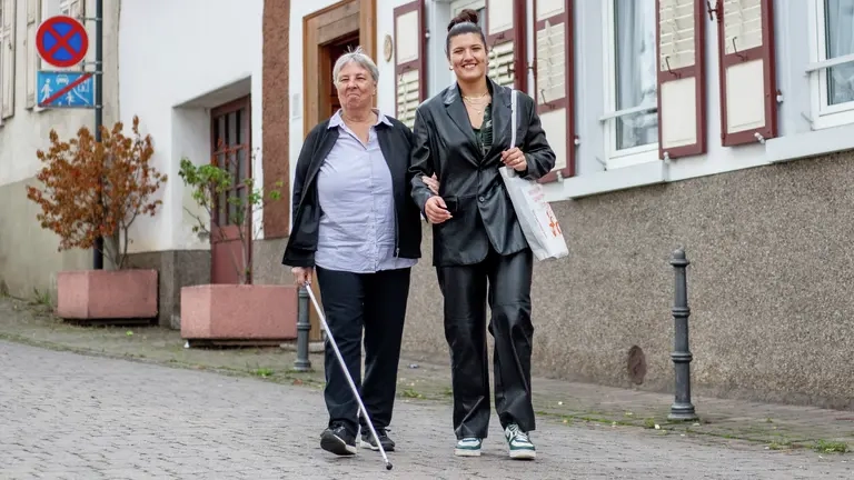 Zwei Frauen gehen die Straße entlang, eine hat einen Blindenstock